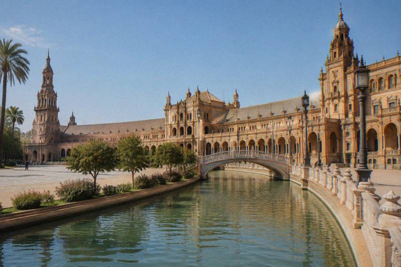 Sunny late-morning view of the vast semicircular brick Plaza de Espana in Seville from across the water channel, the curved colonnade and its twin towers, a small arched bridge with tiled balustrade, a few tiny distant figures far off.