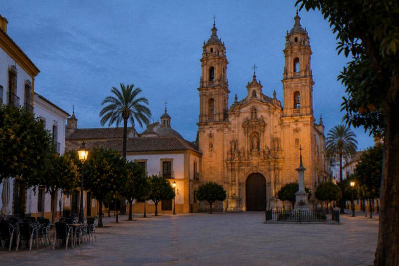Hora azul en la plaza frente a la iglesia barroca del Salvador en Sevilla, su fachada de piedra cálida y sus dos torres iluminadas suavemente, plaza vacía con algunas sillas de terraza apiladas y sin gente.