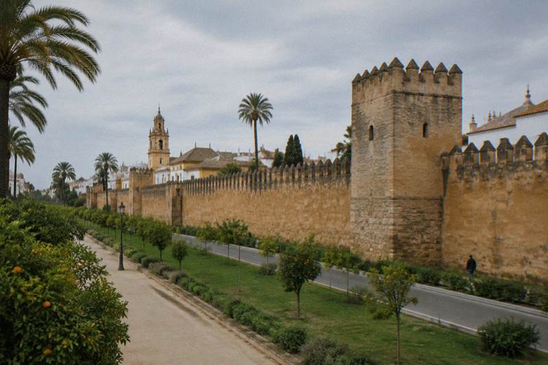 Mañana primaveral nublada en el tramo de la Macarena de la muralla de la ciudad de Sevilla, una larga muralla almenada de color ocre y marrón, con una torre cuadrada, hierba y una calzada a sus pies, y pequeños paseantes a lo lejos.