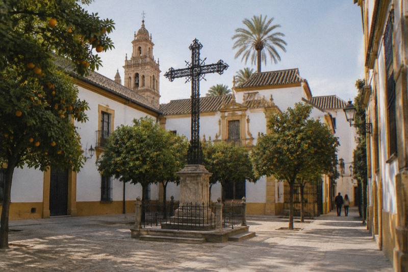 Luz de media mañana en la pequeña plaza de Santa Cruz en Sevilla, con una cruz alta de hierro forjado negro sobre una base de piedra en el centro, naranjos, paredes encaladas con bordes ocre y una pareja diminuta a lo lejos de espaldas.