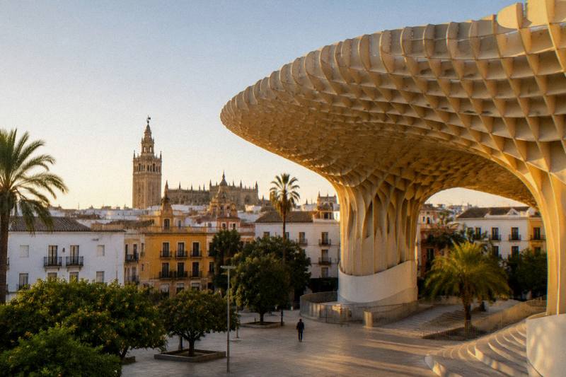 Golden hour at the Metropol Parasol in Seville, the giant undulating wooden mushroom-shaped canopy standing over a square of old low buildings, a plaza with a few tiny distant figures, a clear pale sky.