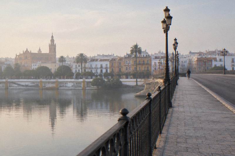 Niebla matutina vista desde el pavimento del puente de Isabel II sobre el Guadalquivir en Sevilla, con barandilla de hierro negro y farolas en primer plano, el río plano y plateado, la torre de la Giralda y las casas ribereñas al otro lado del agua, un ciclista lejano de espaldas.
