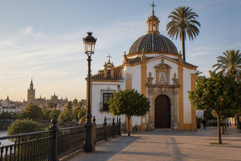 Late-afternoon light on the small whitewashed Chapel of Carmen at the Triana end of the bridge in Seville, ochre and white walls with a tile-decked dome, wrought-iron railings and a street lamp, empty pavement.