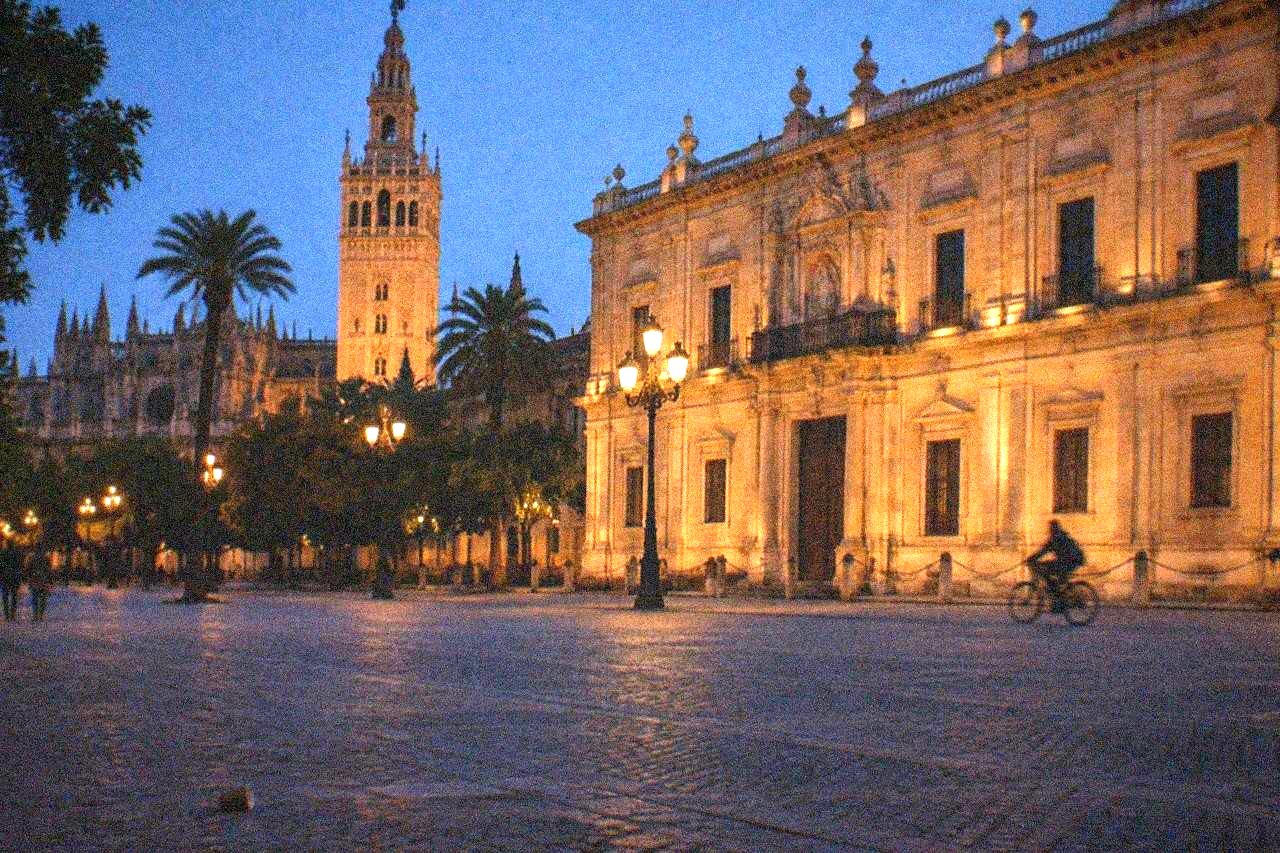 Illuminated stone buildings around an empty evening square in Seville