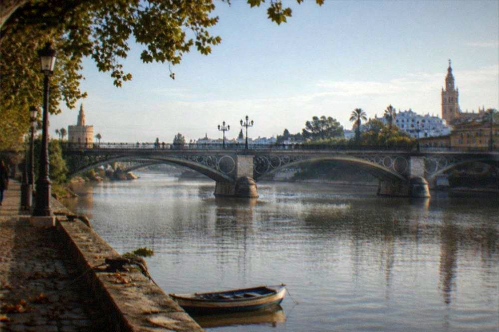 The Triana bridge arching over the Guadalquivir River with the riverbank promenade below