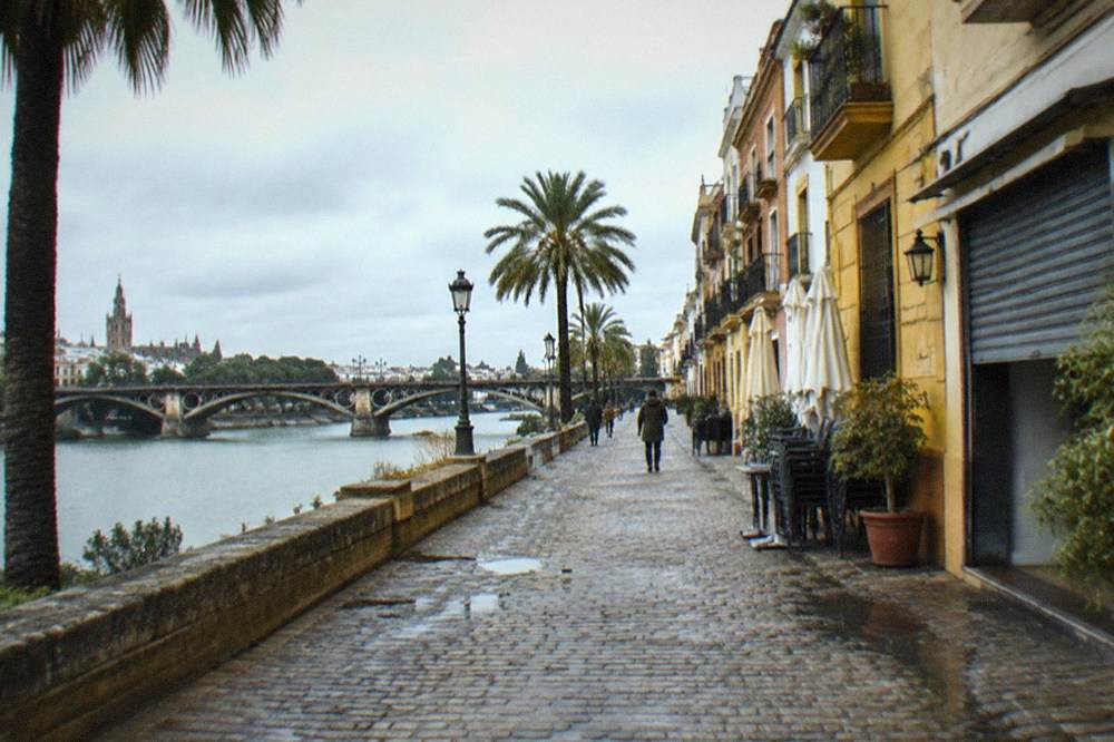 Calle ribereña en Triana con fachadas pastel y un puente de piedra sobre el Guadalquivir