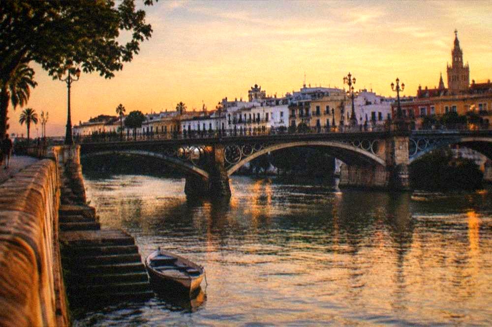 River bank at golden hour with a stone bridge crossing to a low quarter of tiled facades