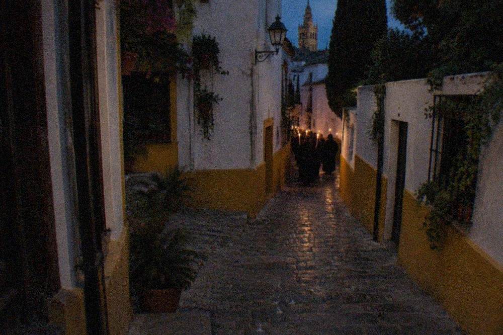 A narrow Seville street at night lit by candles ahead of a procession.