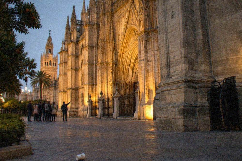 A guide speaking to a small group beside a lit Seville cathedral facade in the evening.
