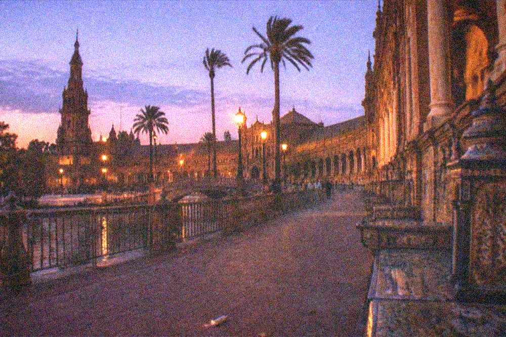 A wide Seville plaza at dusk with palm trees and crowds gathered along the barriers.